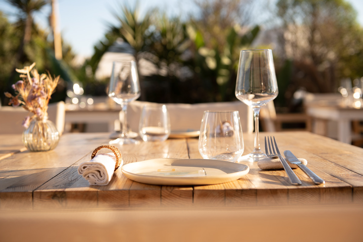Sunset Antiparos | Restaurant in Antiparos, Greece | Photograph of a table setting with plate, glasses, and silverware in the foreground and a sunny out of focus leafy background