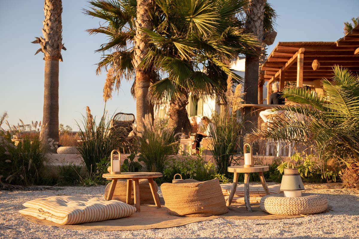 Sunset Antiparos | Restaurant in Antiparos, Greece | Photograph of a seating area on the beach sand of Antiparos, Greece for Sunset Antiparos restaurant. Palm trees in the background