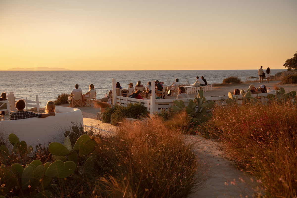 Sunset Antiparos | Restaurant in Antiparos, Greece | Photograph with bushy foliage in foreground and restaurant goers sitting in the background looking out at the ocean